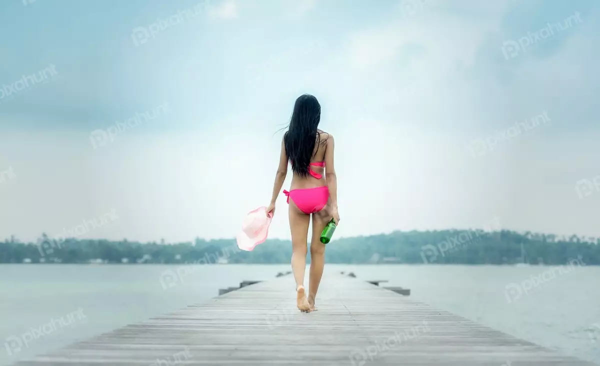 Free Premium Stock Photos A girl in a pink bikini walking away from the camera on a wooden dock and long dark hair is blowing in the wind and she is carrying a bottle of wine and a hat.