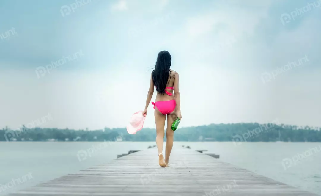 Free Premium Stock Photos A girl in a pink bikini walking away from the camera on a wooden dock and long dark hair is blowing in the wind and she is carrying a bottle of wine and a hat.